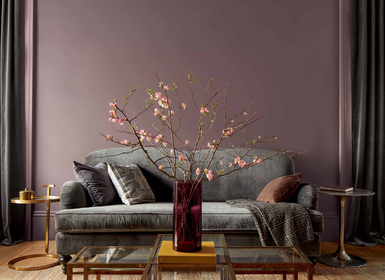 A purple-painted living room features a gray couch, gold side tables, and a glass-topped gold coffee table topped with a red vase filled with pink flowers.