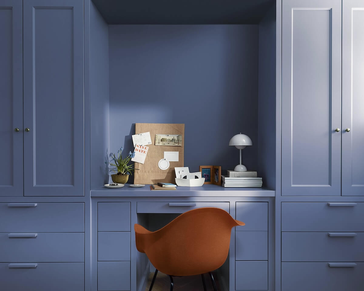 A modern red office chair sits in a home office painted in Blue Nova 825, with built-in shelving, cabinetry and in-set desk topped with a pegboard, plants, books and a modern domed white desk lamp.