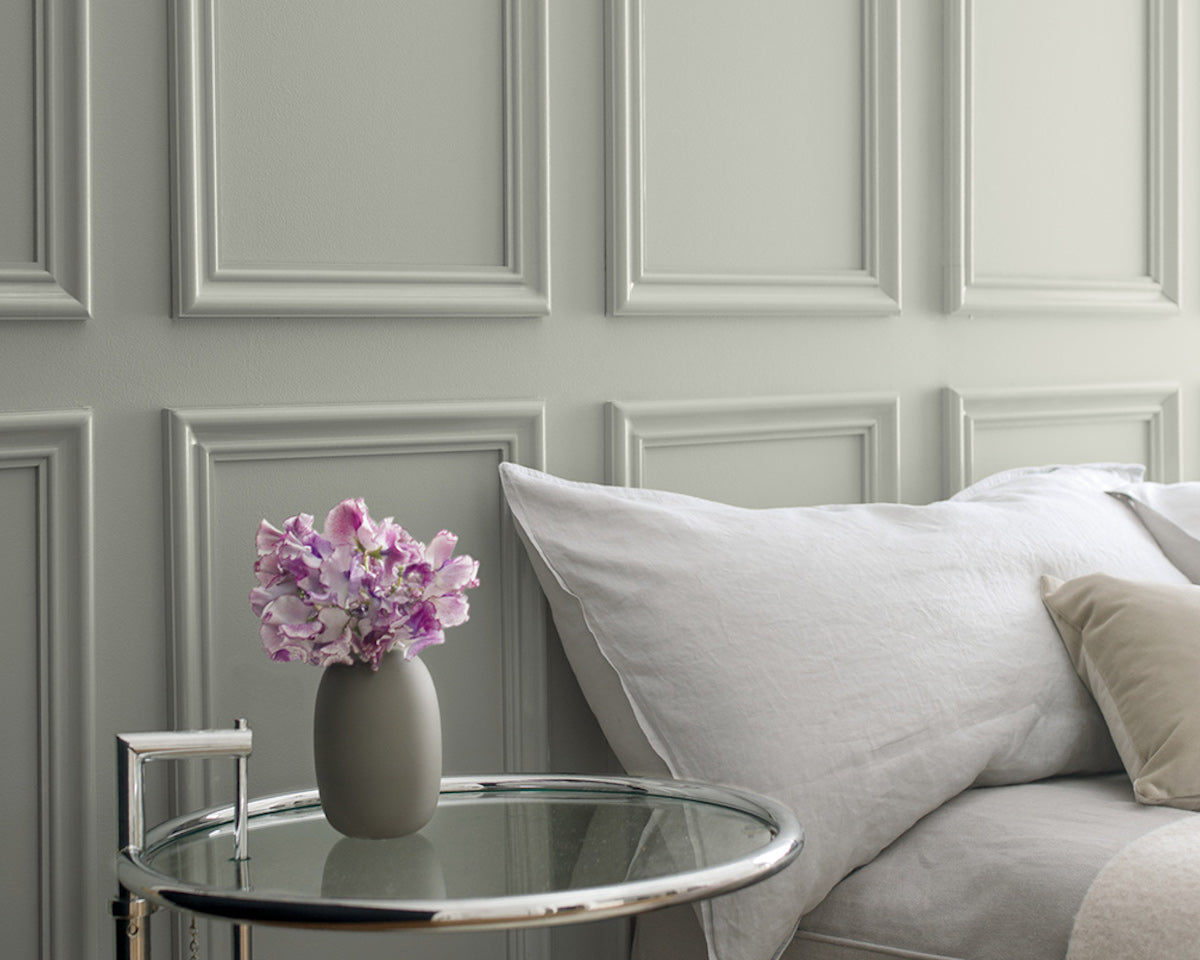 Gray wainscoting behind a bed with gray bedding and a glass side table featuring a small vase of purple flowers.