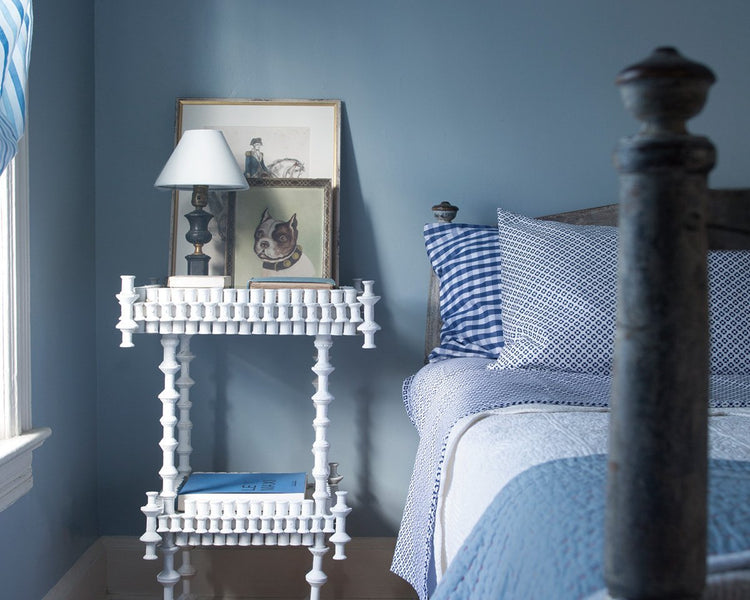 Bedroom with blue walls, small white table with lamp, and bed with blue sheets and pillows beside it